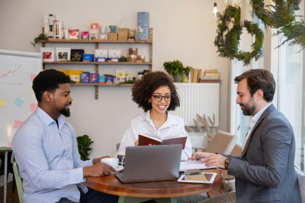 Three people are engaging in a discussion at a wooden table with a laptop, notebooks, and a coffee cup. They are smiling, suggesting a positive interaction.