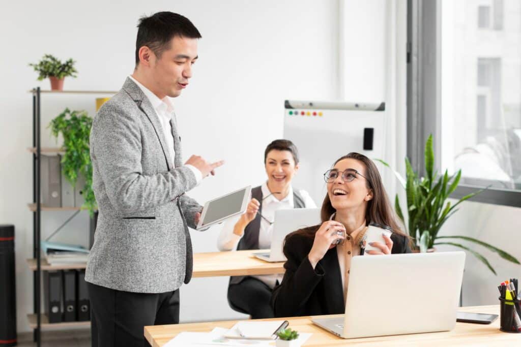 Three professionals in an office setting, one standing with a tablet, two sitting at a desk with laptops, all appear engaged and happy.