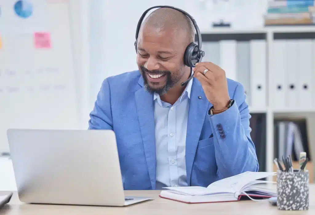 A person in a blue suit is smiling while working on a laptop, wearing a headset with a microphone, possibly engaged in a video call.